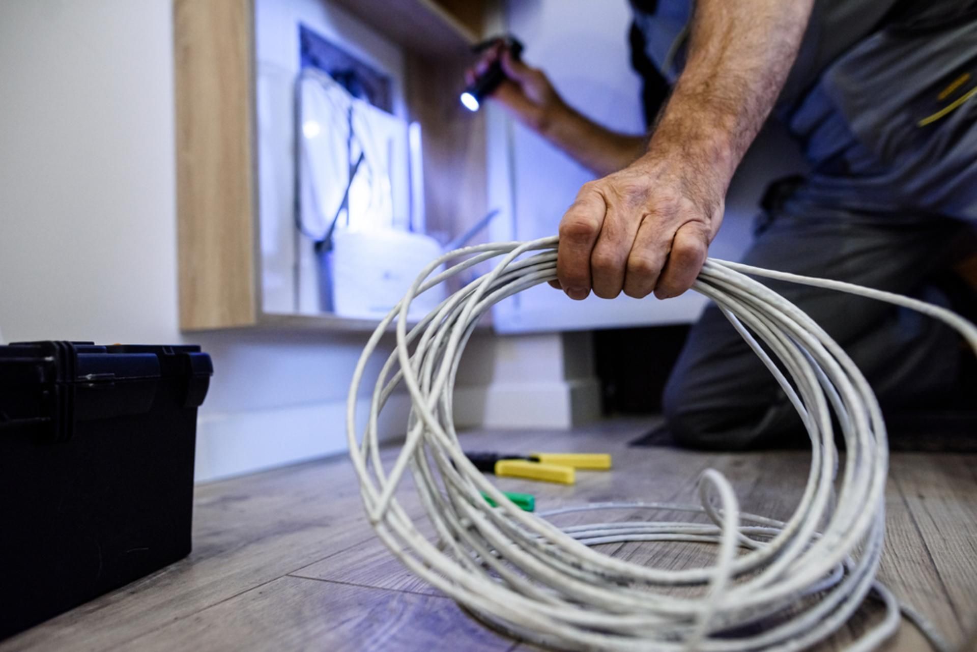 Electrician holding a coil of wire, inspecting a cabinet with a flashlight, inside a home.