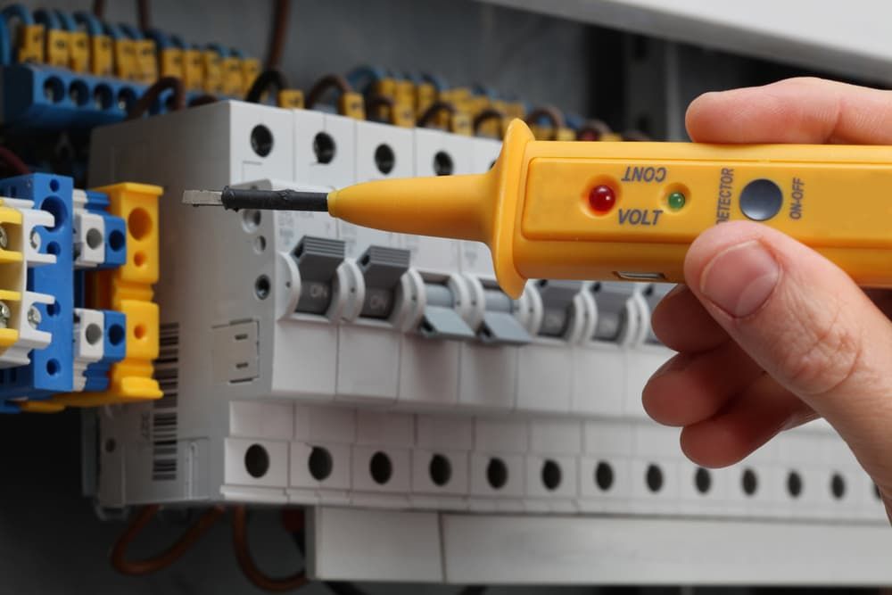 A Hand Holds a Yellow Voltage Tester Near a Circuit Breaker Panel