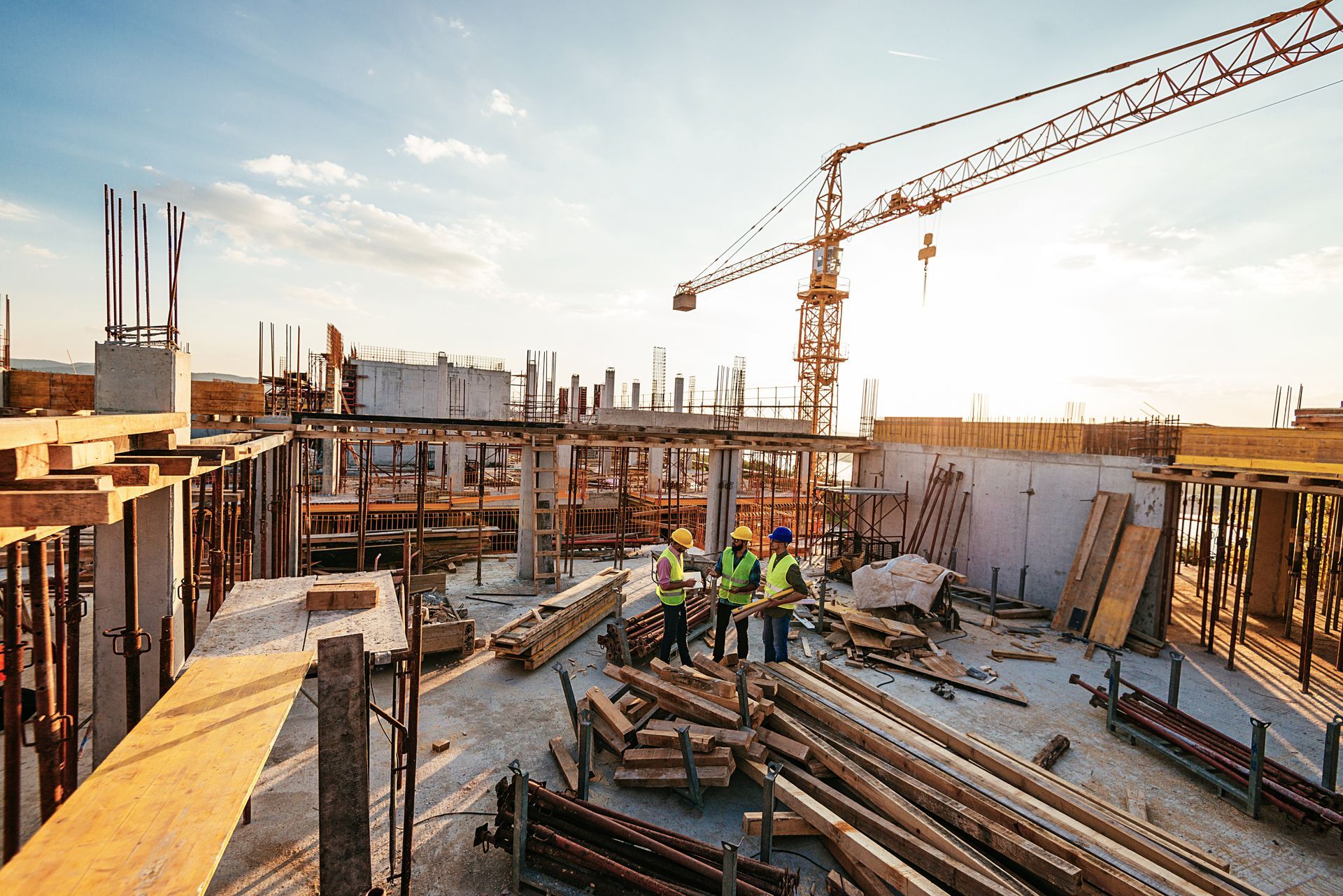 Construction site with crane, workers in safety vests, concrete, wood, and a bright sky.