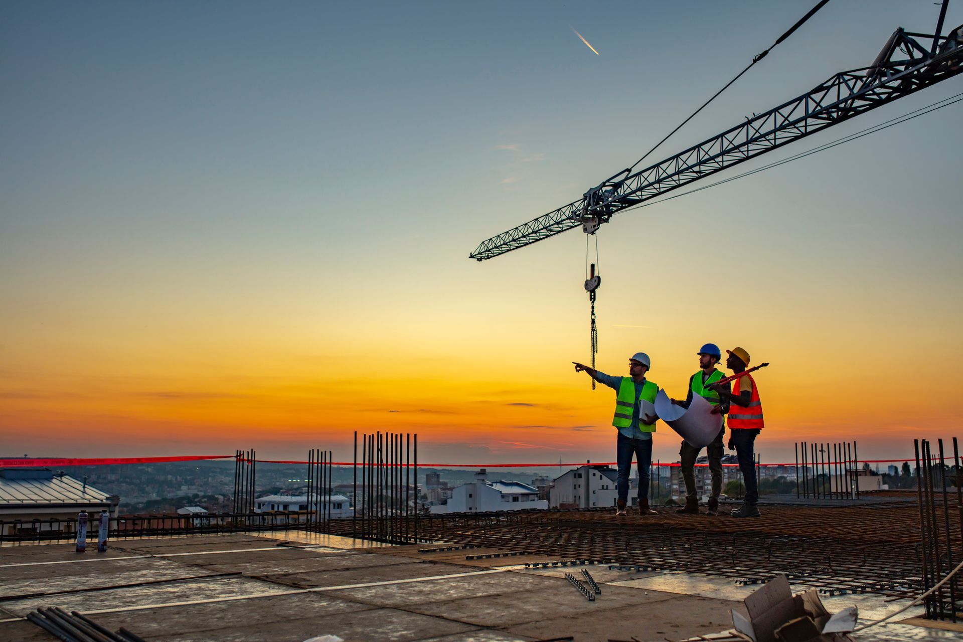 Construction workers on a building site, gesturing at plans, with a crane and sunset.