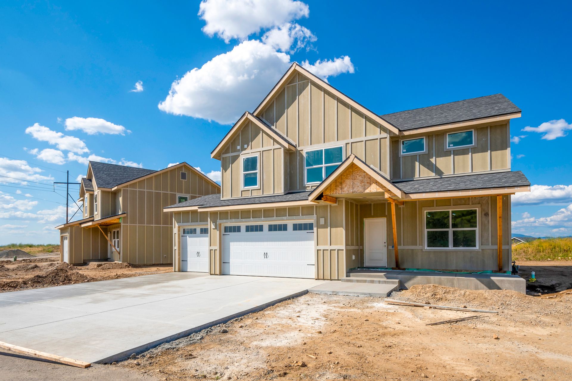 New construction two-story house with tan siding, a white garage door, and a concrete driveway under a blue sky.