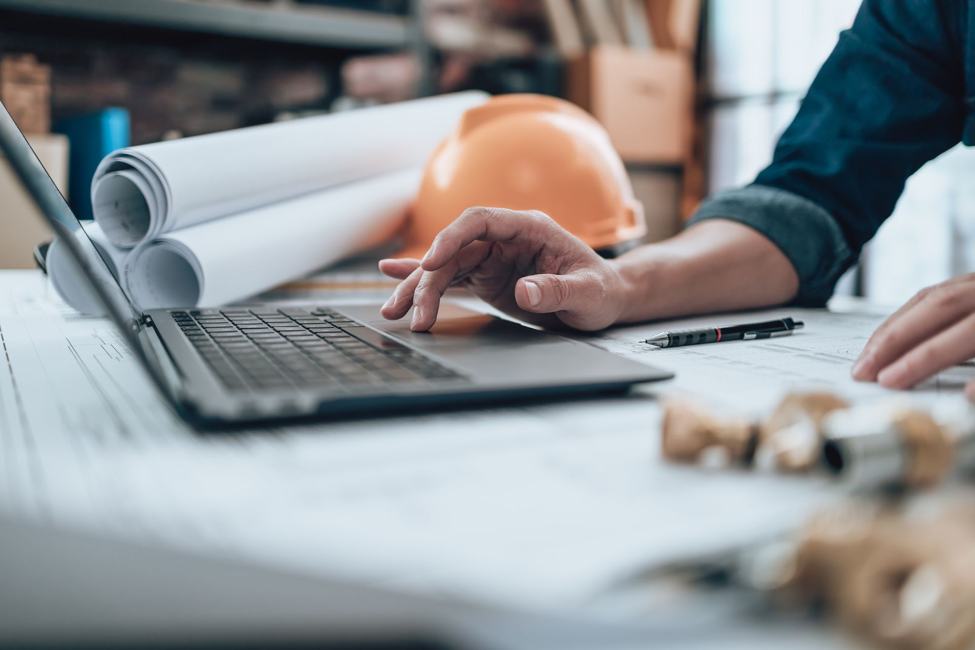 Person working on laptop at a desk with blueprints, a hard hat, and rolled documents.
