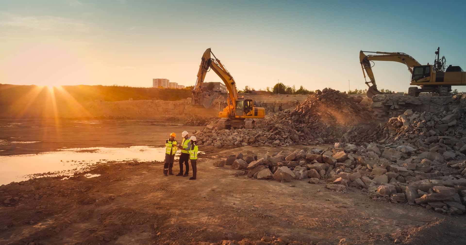 Construction site at sunset with workers in safety vests, excavators, and piles of rocks.