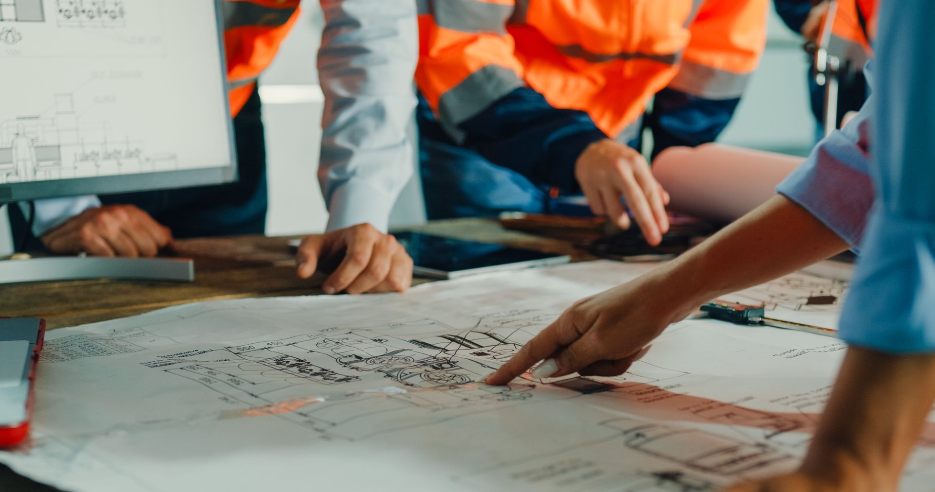 Construction workers reviewing blueprints, pointing at details on a table with a computer monitor.