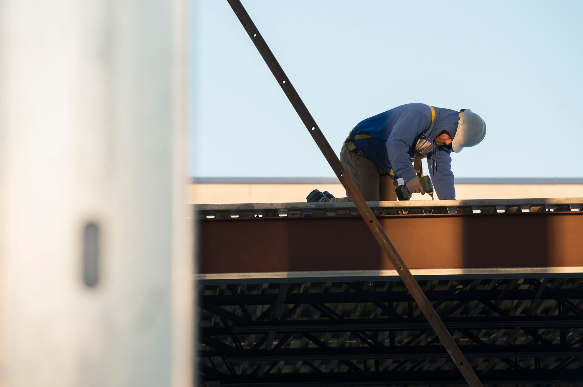 Person on a roof working with a drill, wearing safety gear. Steel support beam visible.
