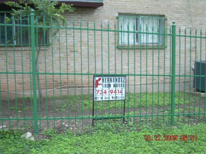 Green metal fence in front of a brick house with a window and a small sign on the gate.