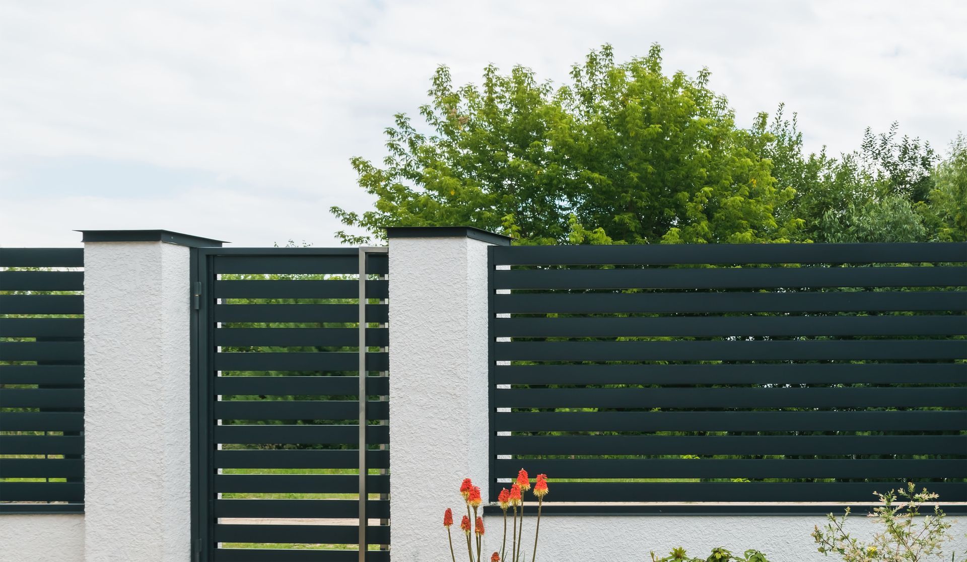 Modern black slat fence with white pillars and red flowers in front of a green tree