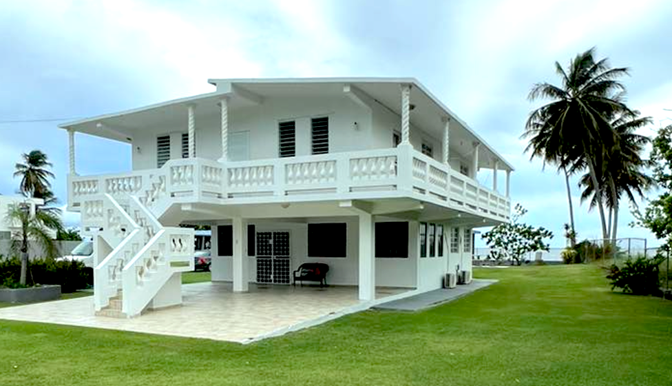 Two-story white house with balcony, stairs, and open ground floor on a grassy lawn near the ocean.