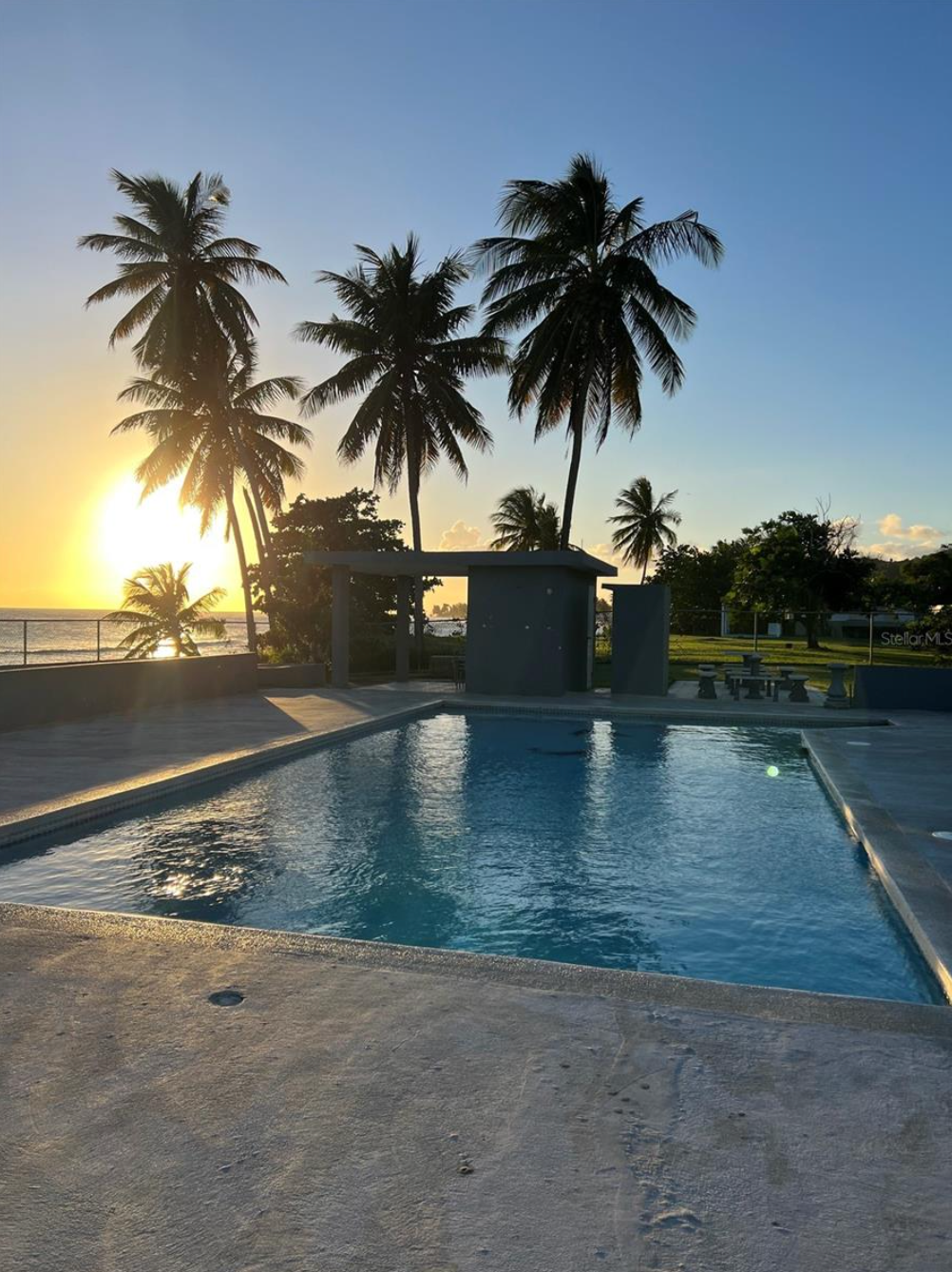 Palm trees frame a pool reflecting a sunset over the ocean.
