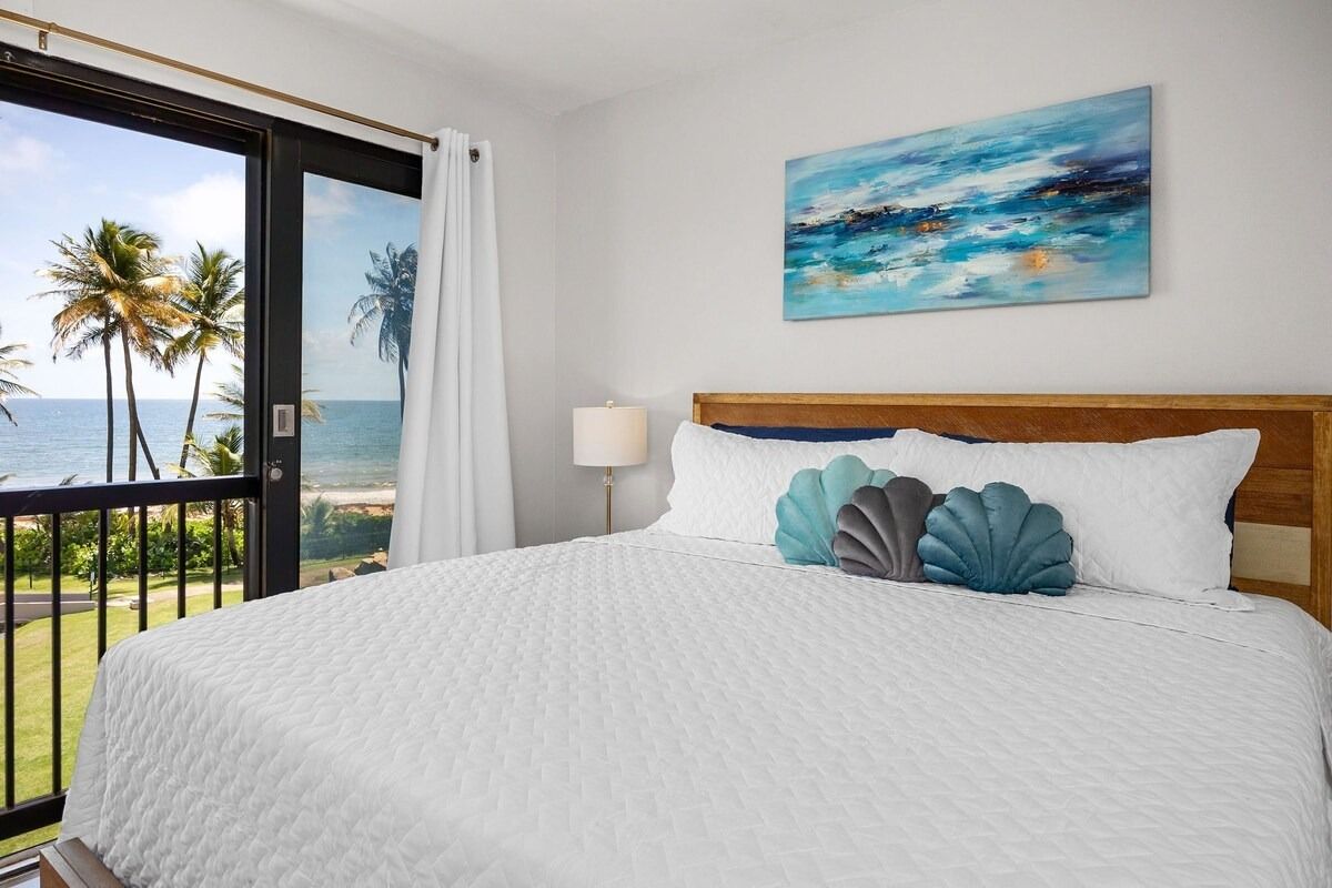 Bedroom with ocean view; bed with white comforter, shell pillows, artwork, sliding glass door, and balcony.