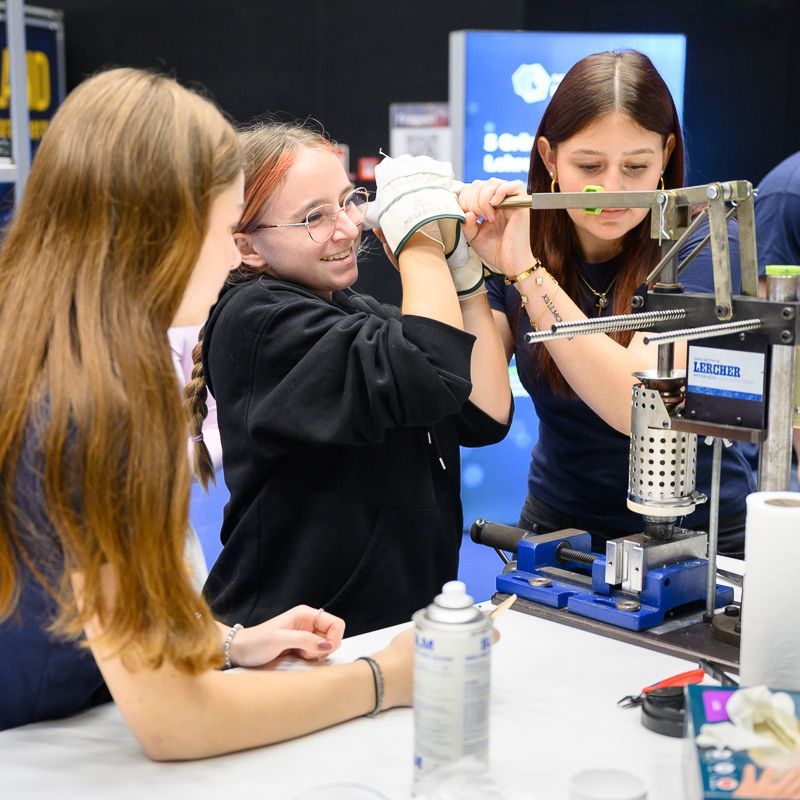 Konzentriert und lächelnd arbeiten zwei Studierende bei einer Robotik-Veranstaltung an einem Roboterarm.