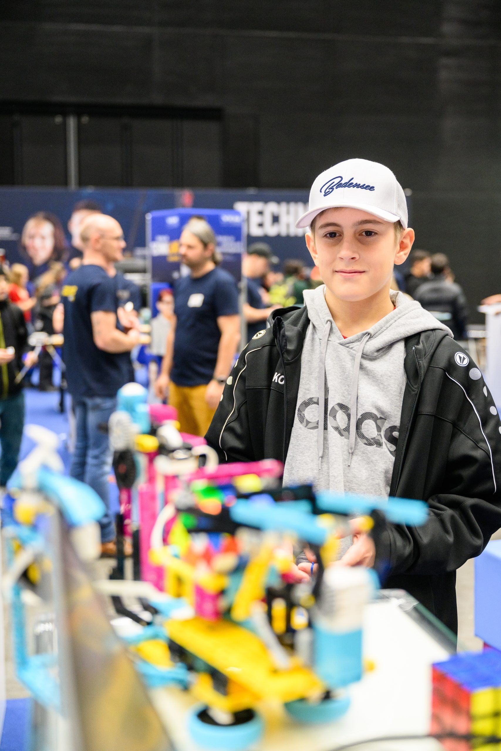 Ein Teenager mit Mütze und Jacke lächelt und steht bei einer Veranstaltung neben einer farbenfrohen Roboterausstellung.