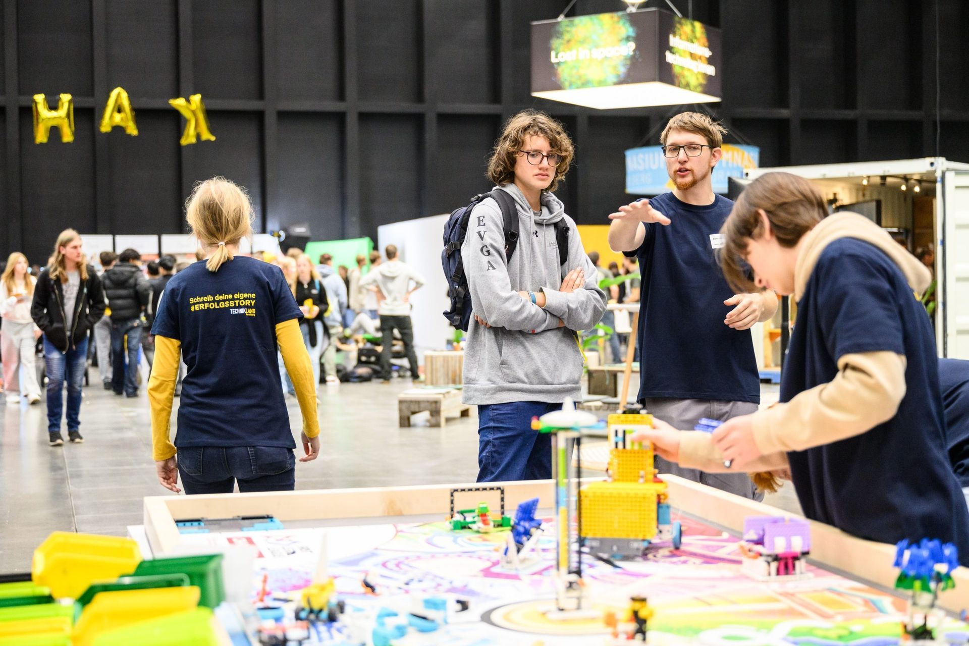 Menschen interagieren auf einer Tagung mit einem farbenfrohen Projekttisch, im Hintergrund sind „HAX“-Ballons zu sehen.