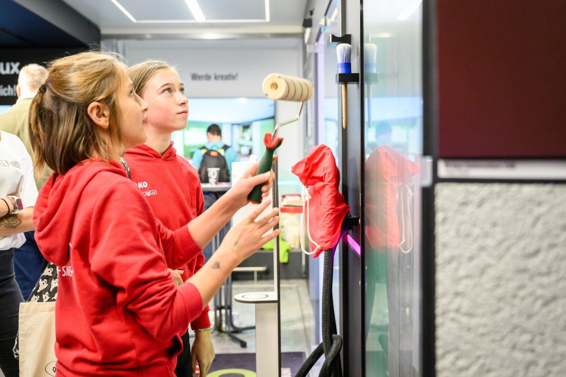 Zwei Mädchen in roten Hemden streichen mit Farbrollen an einer Wand und blicken nach oben. Drinnen.