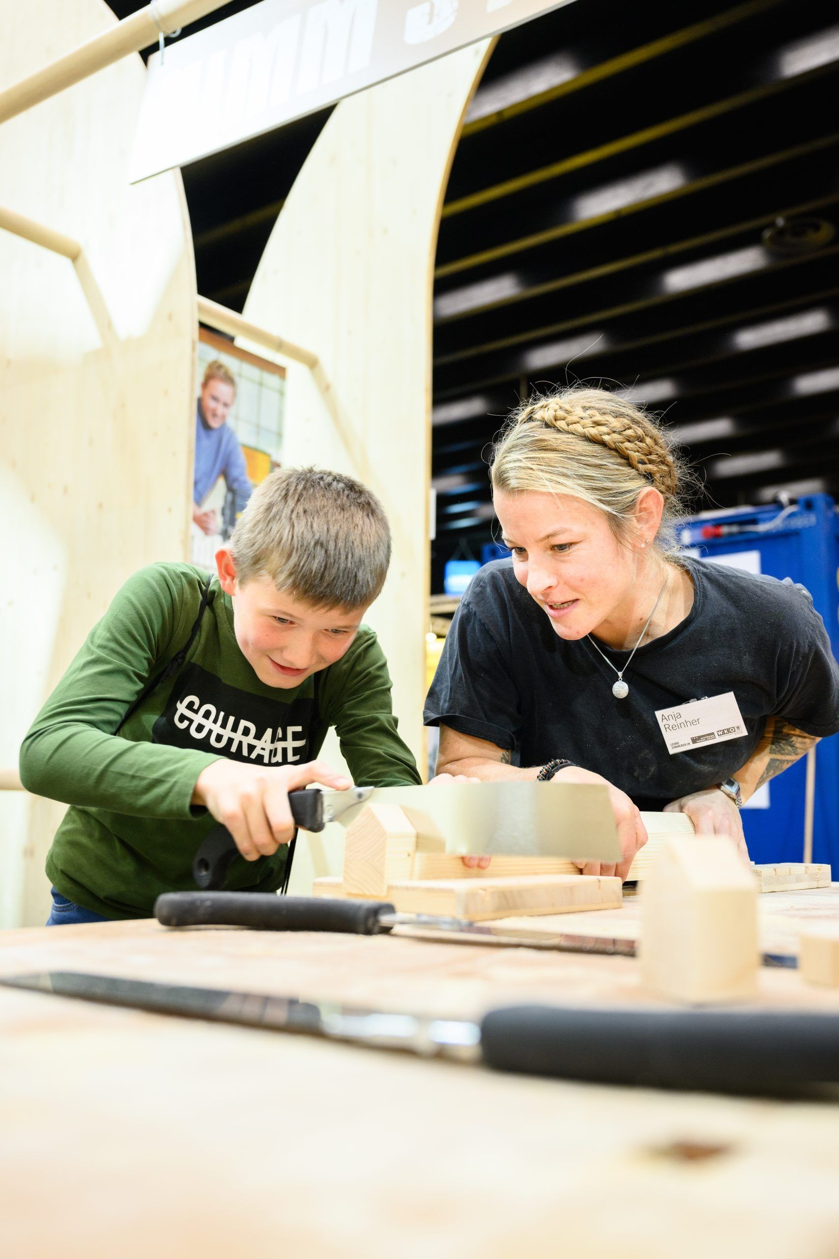 Junge und Frau arbeiten gemeinsam an Holz. Junge sägt Holz, Frau führt. Drinnen, Naturholz, fröhliche Ausdrücke.