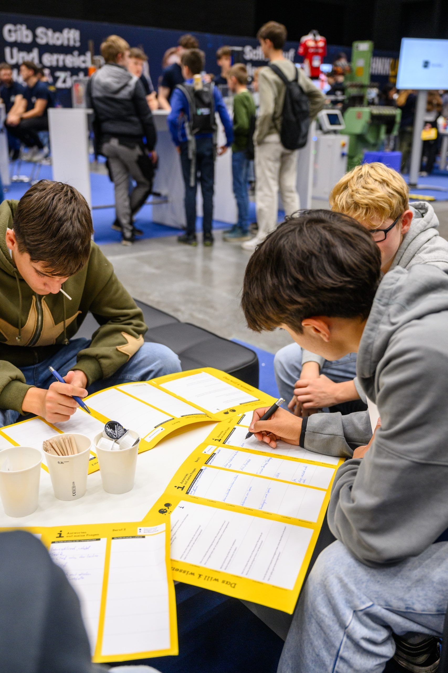 Vier junge Leute füllen bei einer Veranstaltung gelbe Formulare aus, andere im Hintergrund.