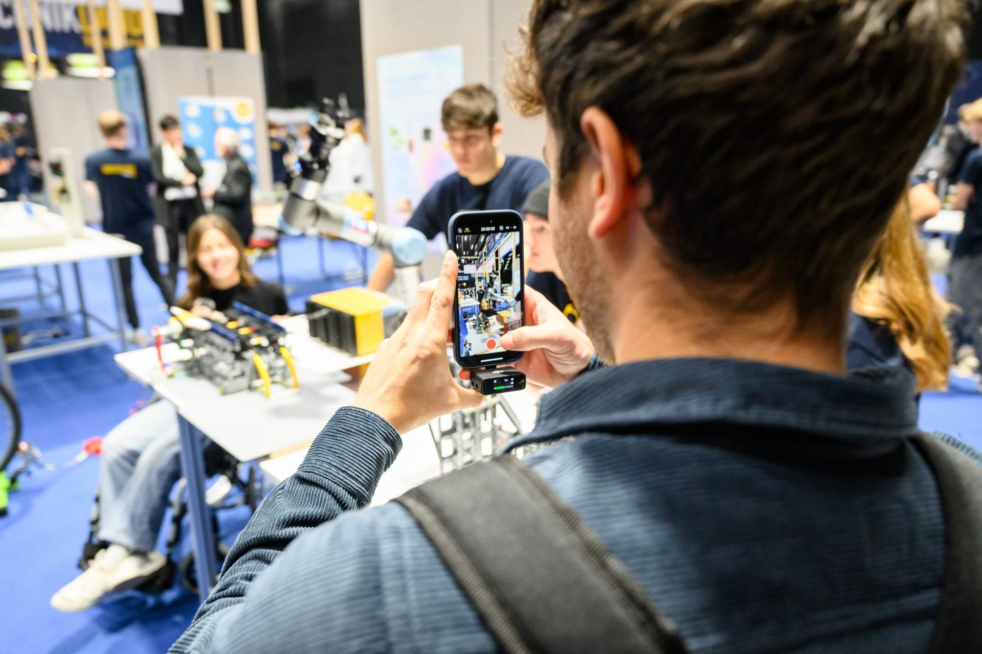 Mann macht ein Foto von einer Roboterausstellung auf einer Konferenz. Im Hintergrund sind Menschen zu sehen.