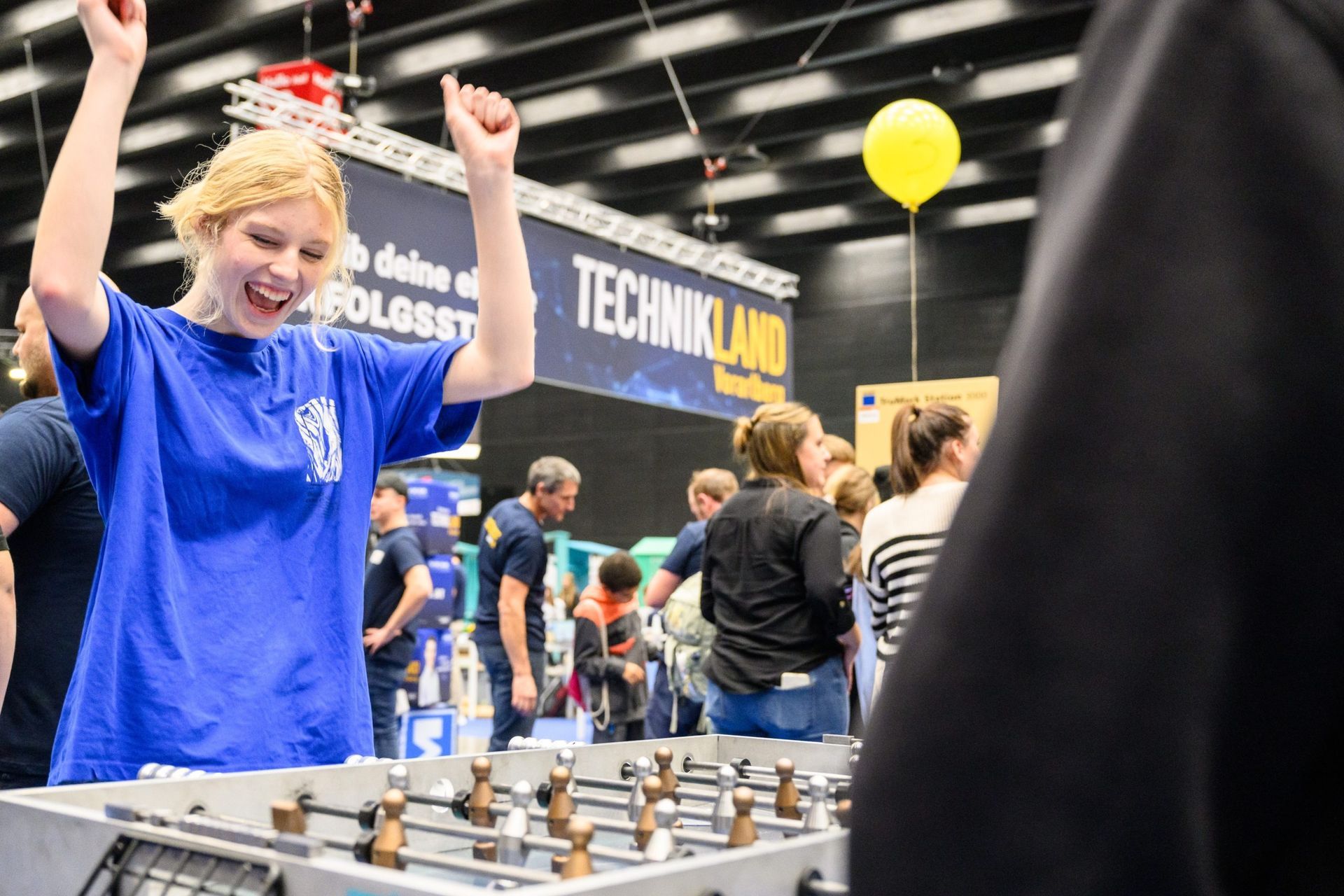 Eine junge Frau in einem blauen Hemd jubelt mit erhobenen Armen beim Tischfußballspielen bei einer Veranstaltung,