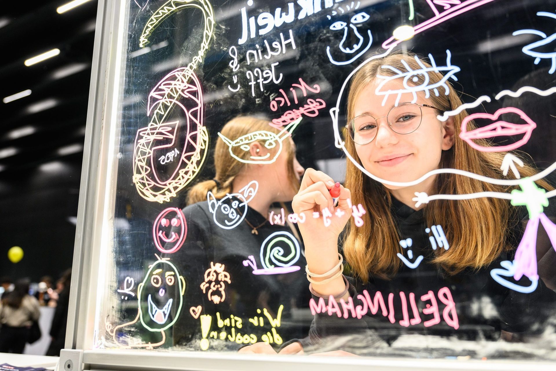 Eine junge Frau mit Brille zeichnet lächelnd bunte Kritzeleien auf Glas, während eine andere Person zusieht.