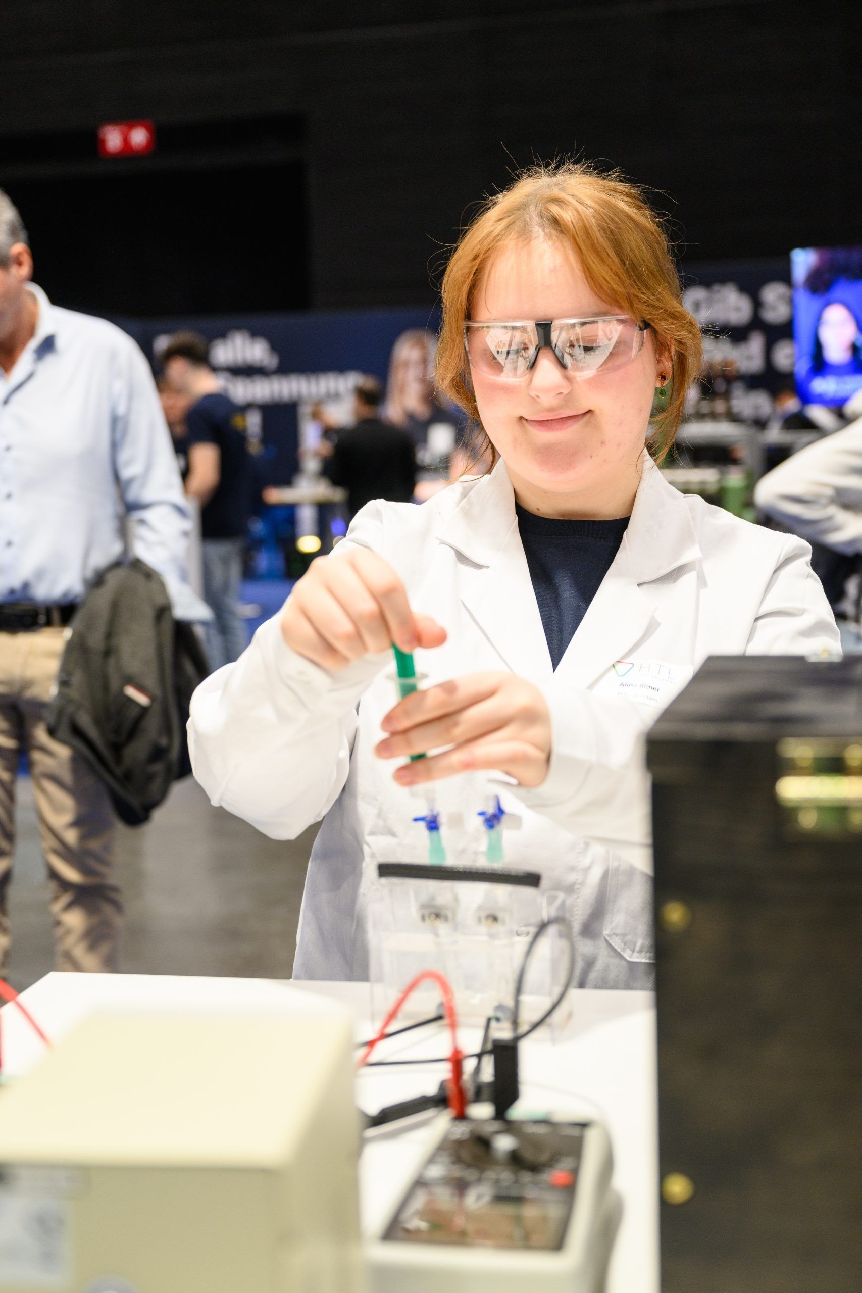 Junge Frau im Laborkittel und mit Schutzbrille lächelt, während sie drinnen an einem wissenschaftlichen Experiment arbeitet.