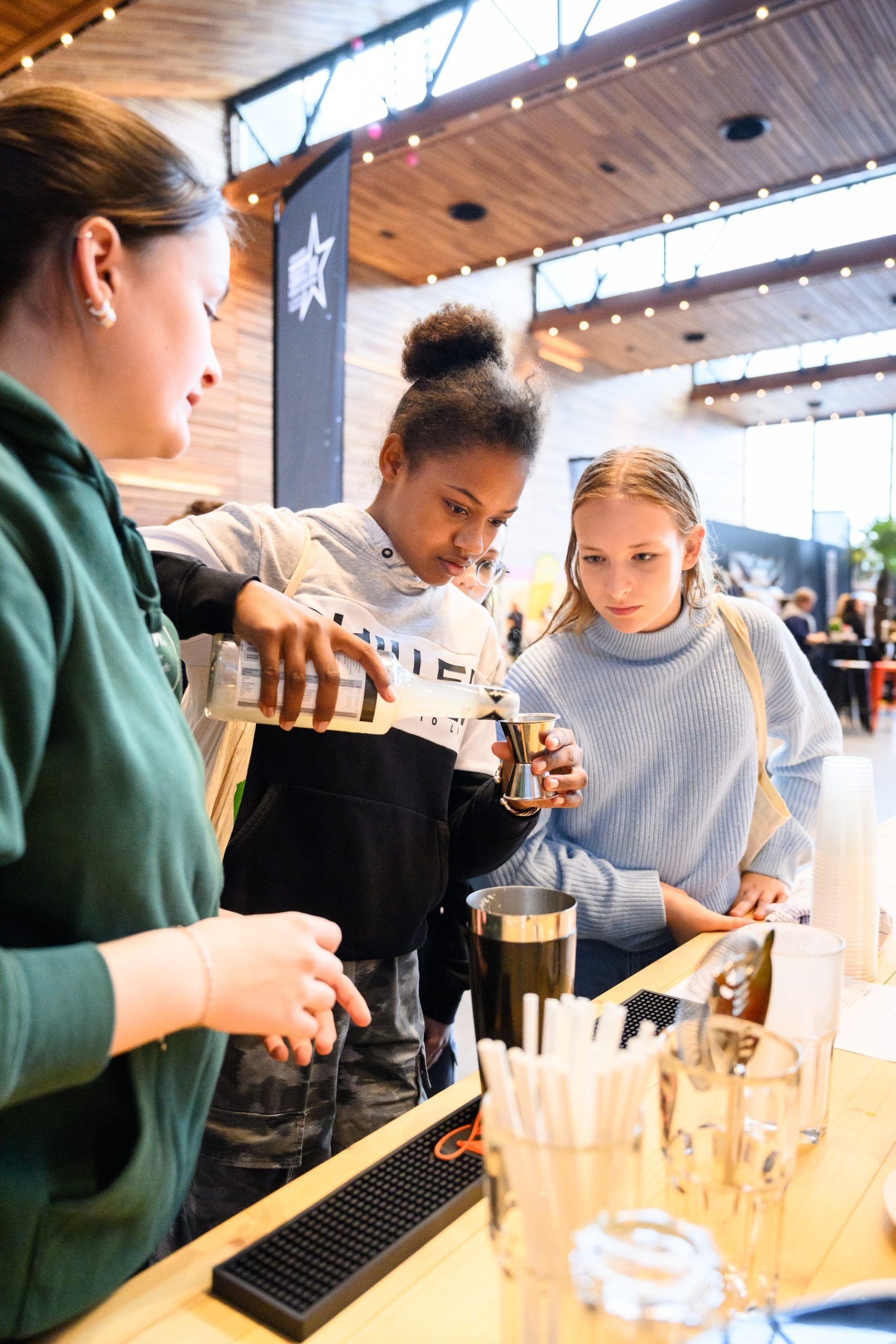 Drei junge Leute bereiten hinter einer Bar einen Drink zu, einer schenkt aus einer Flasche ein.