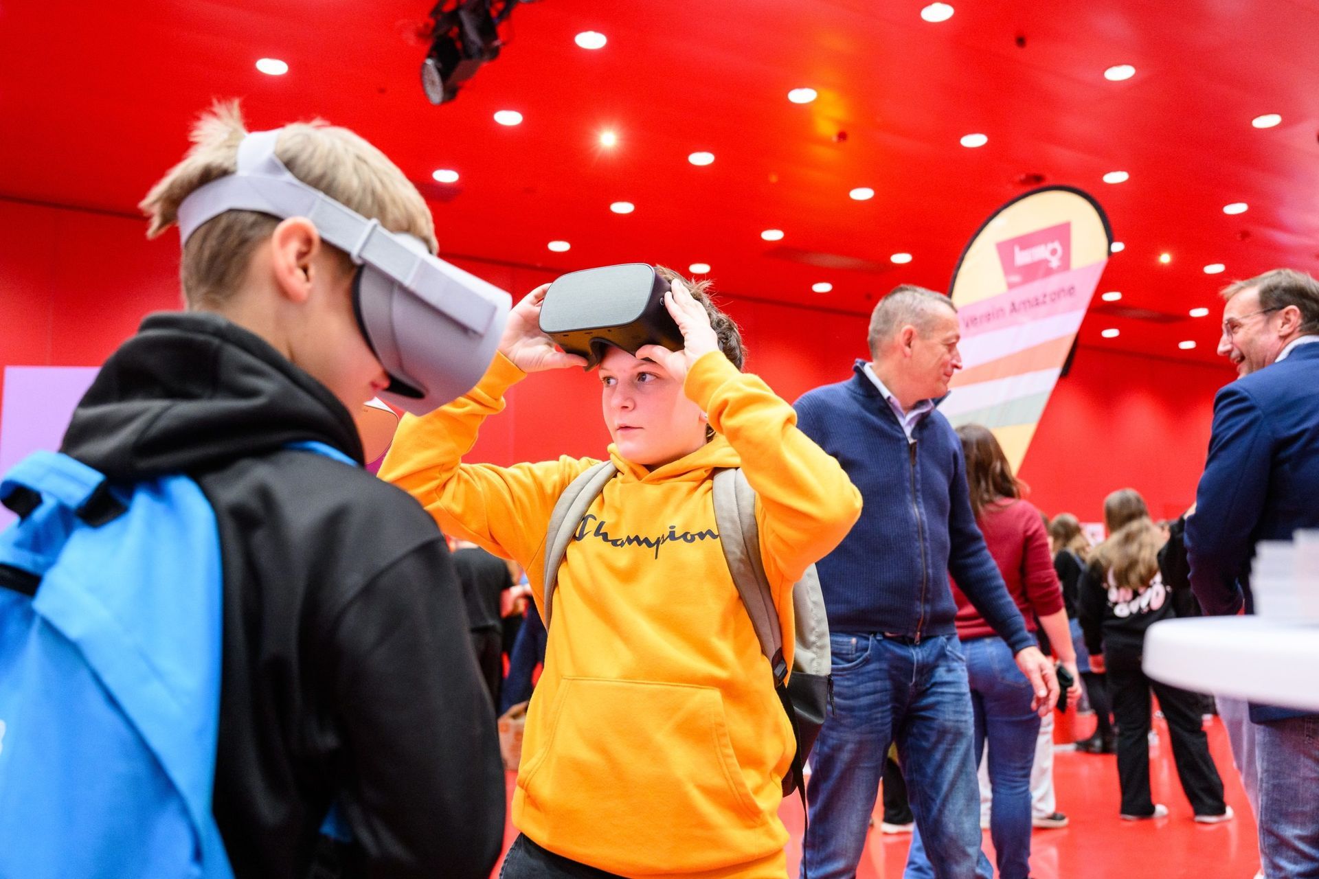 Junge im gelben Kapuzenpulli passt VR-Headset in einem roten Raum an, andere schauen zu.