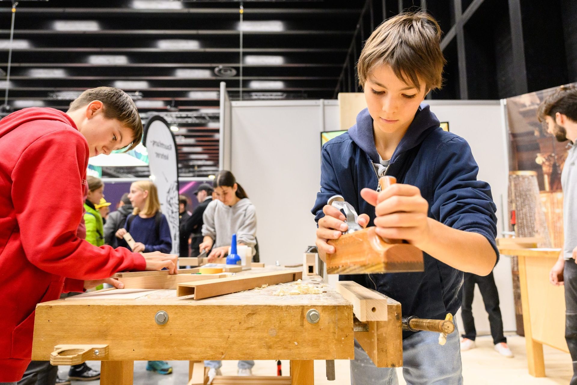 Zwei Jungen verwenden Holzbearbeitungswerkzeuge, einer hobelt ein Holzbrett in einer Werkstatt.