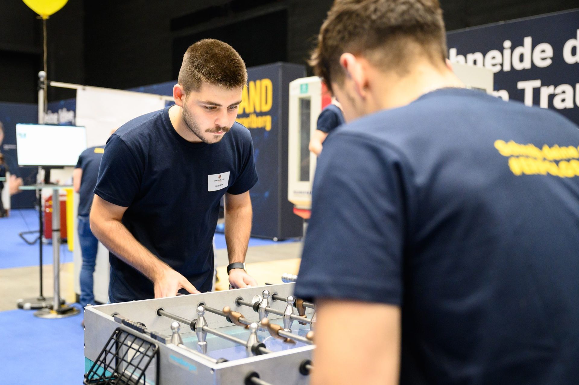 Zwei Männer spielen auf einer Messe Tischfußball. Einer ist konzentriert, der andere schaut nach unten.