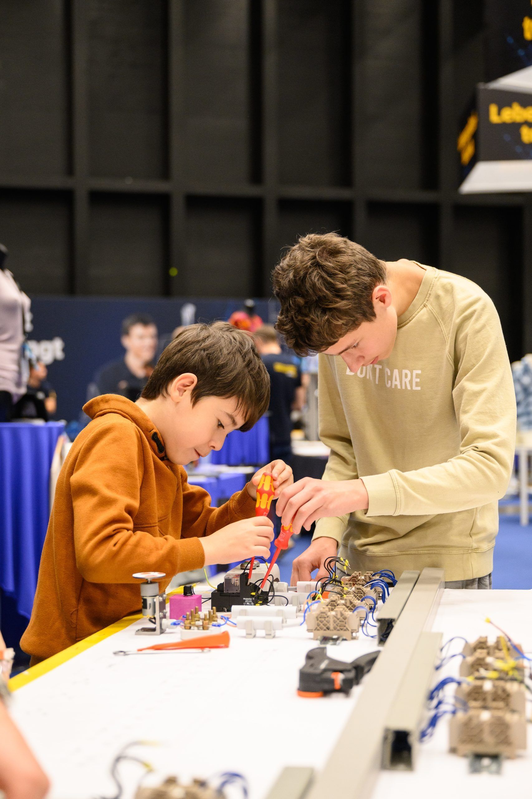 Zwei Jungen bauen an einem Tisch in einer Ausstellungshalle einen Roboter. Einer benutzt einen Schraubenzieher.
