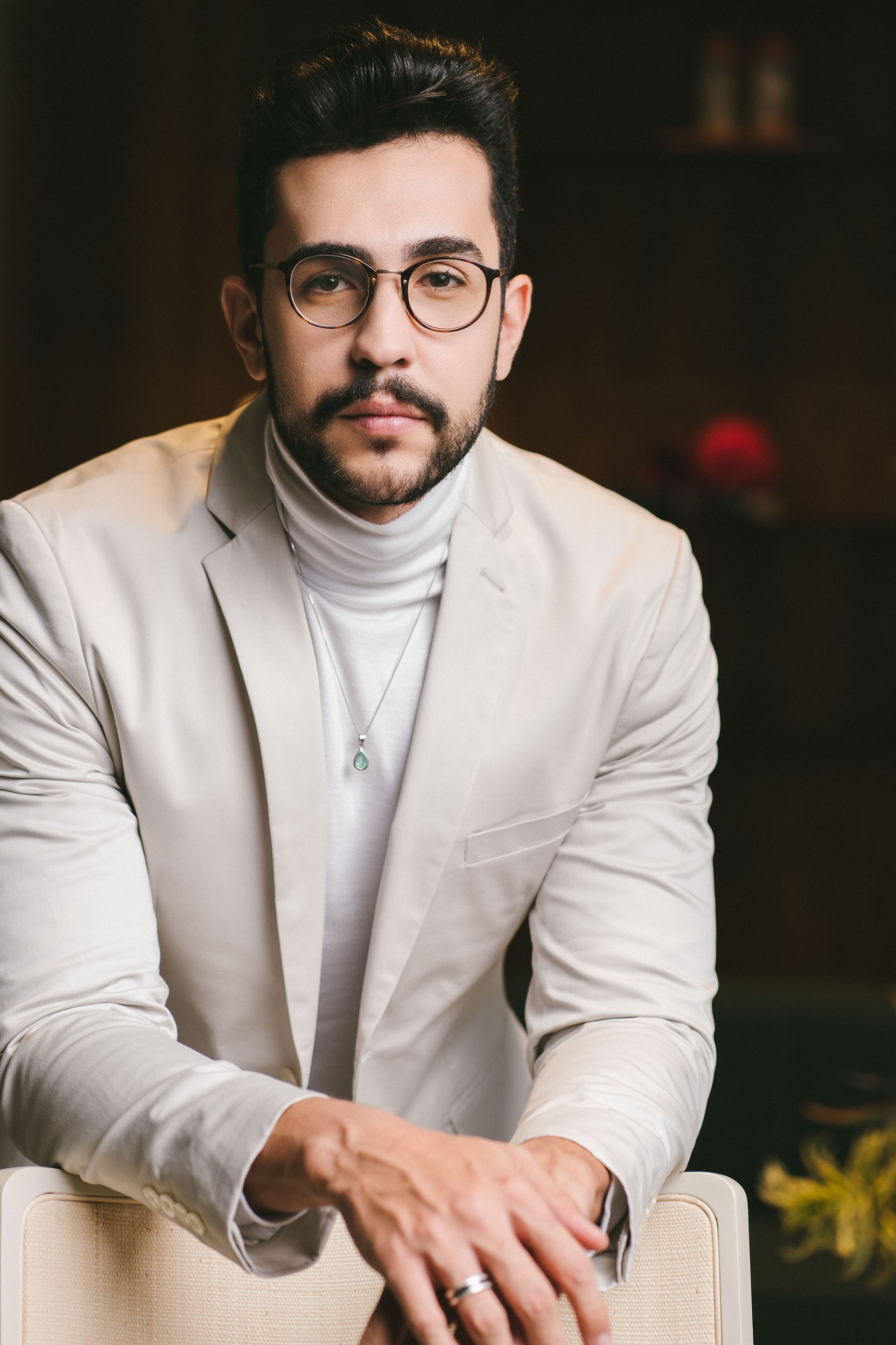 Um homem de óculos e jaqueta branca está sentado em uma cadeira.