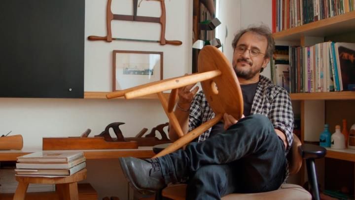 Um homem está sentado em uma cadeira com as pernas cruzadas, segurando uma cadeira de madeira.