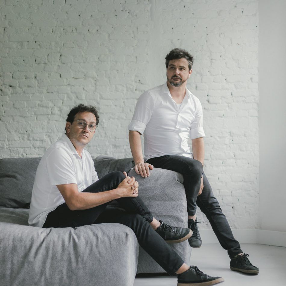 Dois homens estão sentados em um sofá em frente a uma parede de tijolos