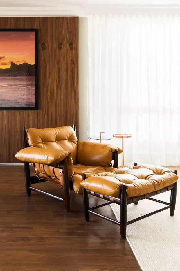 Uma sala de estar com cadeira e pufe e uma pintura na parede.