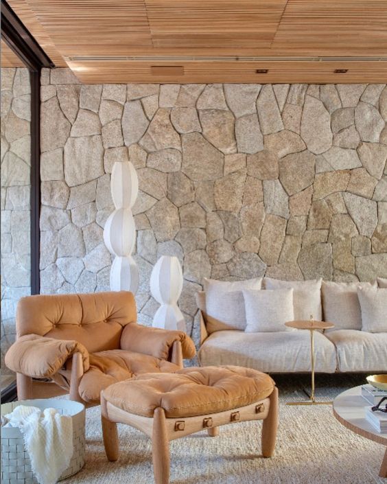 Uma sala de estar com sofá, cadeira e pufe em frente a uma parede de pedra.