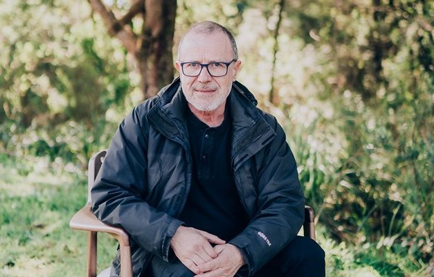 Um homem de jaqueta preta e óculos está sentado em uma cadeira.