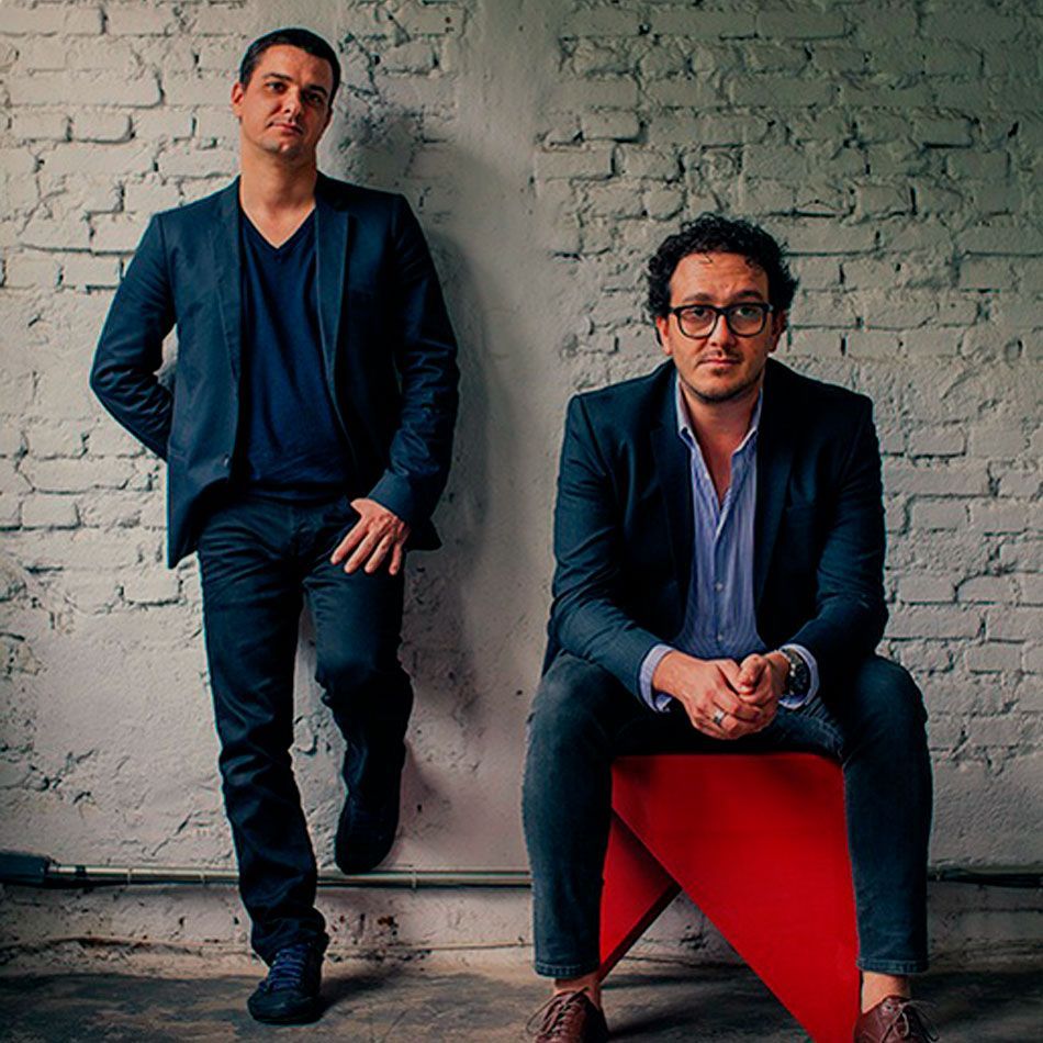 Dois homens de terno estão parados um ao lado do outro em frente a uma parede de tijolos.