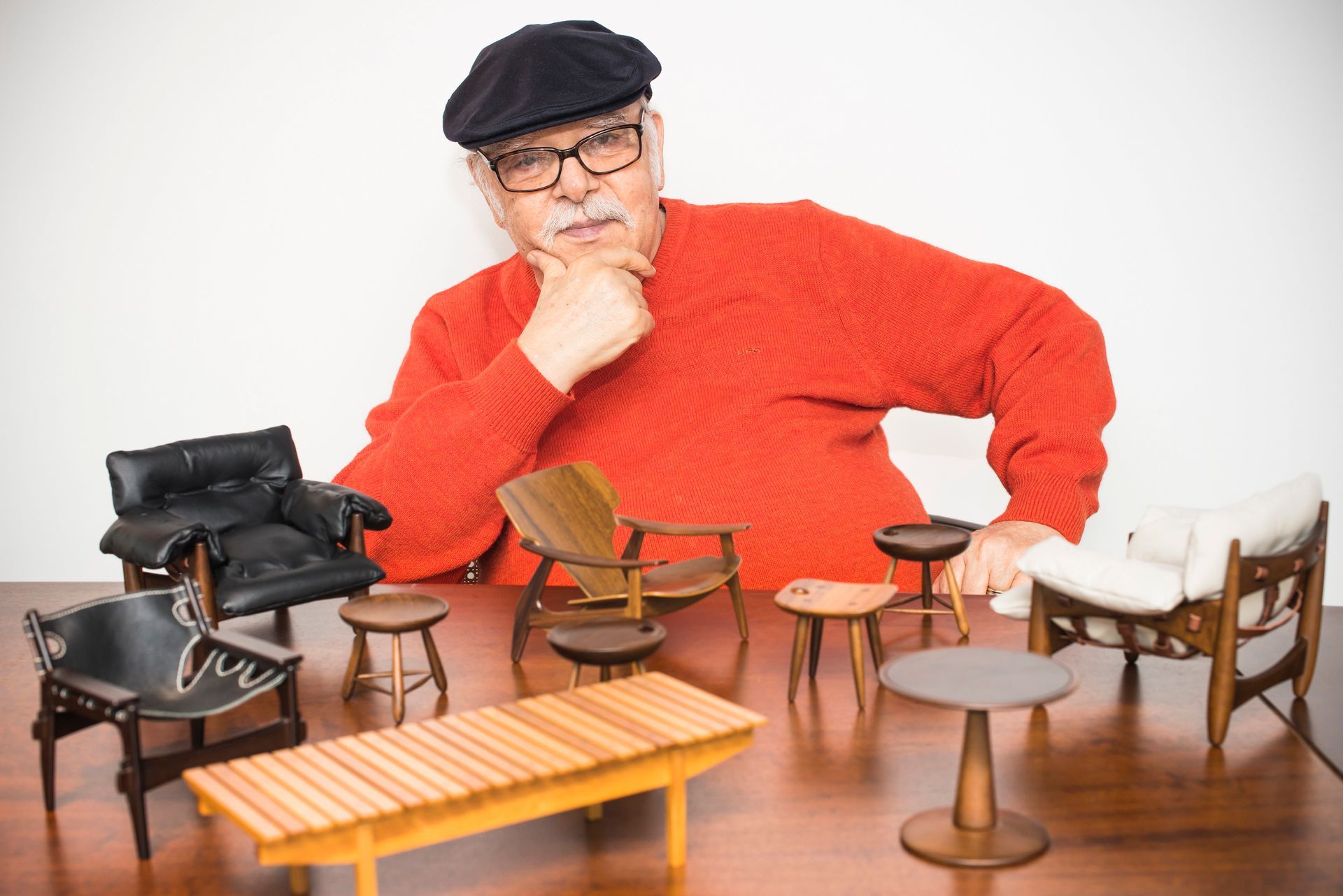 Um homem de suéter vermelho está sentado a uma mesa com móveis em miniatura.