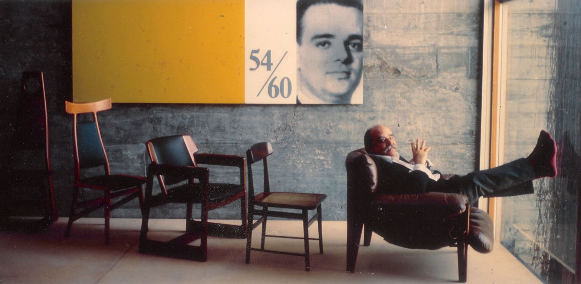 Um homem sentado em uma cadeira com os pés levantados em frente a uma pintura que diz 54/60