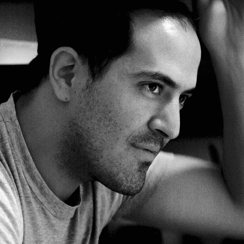 Um homem com barba e camisa cinza está olhando para a câmera em uma foto em preto e branco.