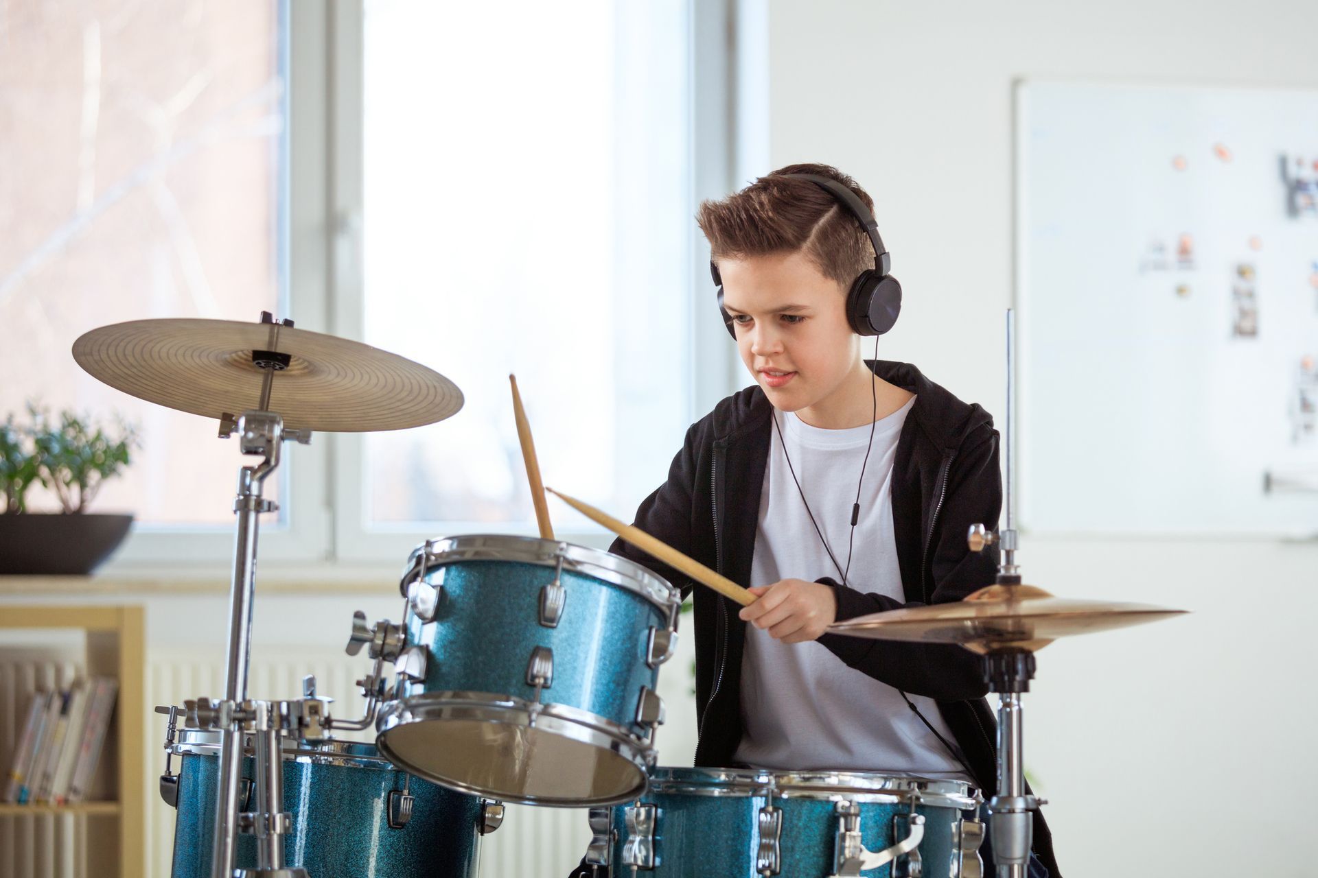 Un ragazzo con le cuffie suona una batteria color verde acqua in una stanza, con le bacchette alzate e un'espressione concentrata.