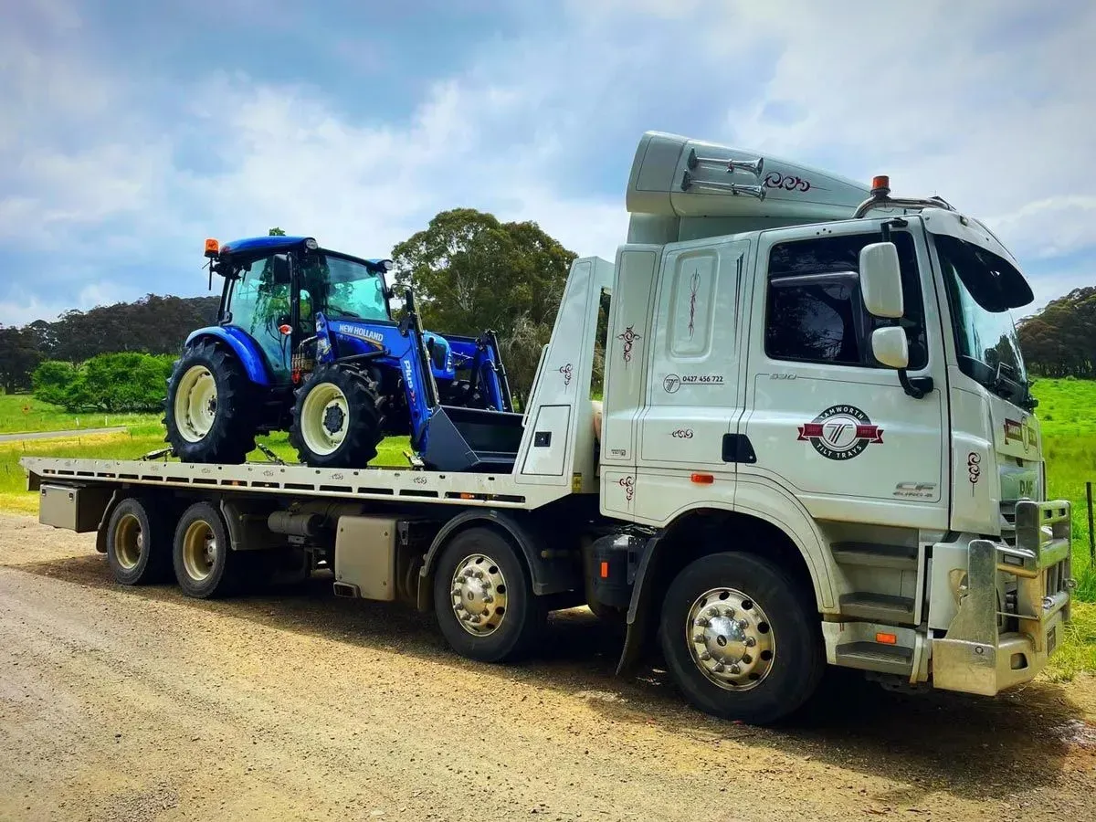A White Flatbed Tow Truck Transporting a Blue New Holland Tractor — Tamworth Tilt Trays in Tamworth, NSW