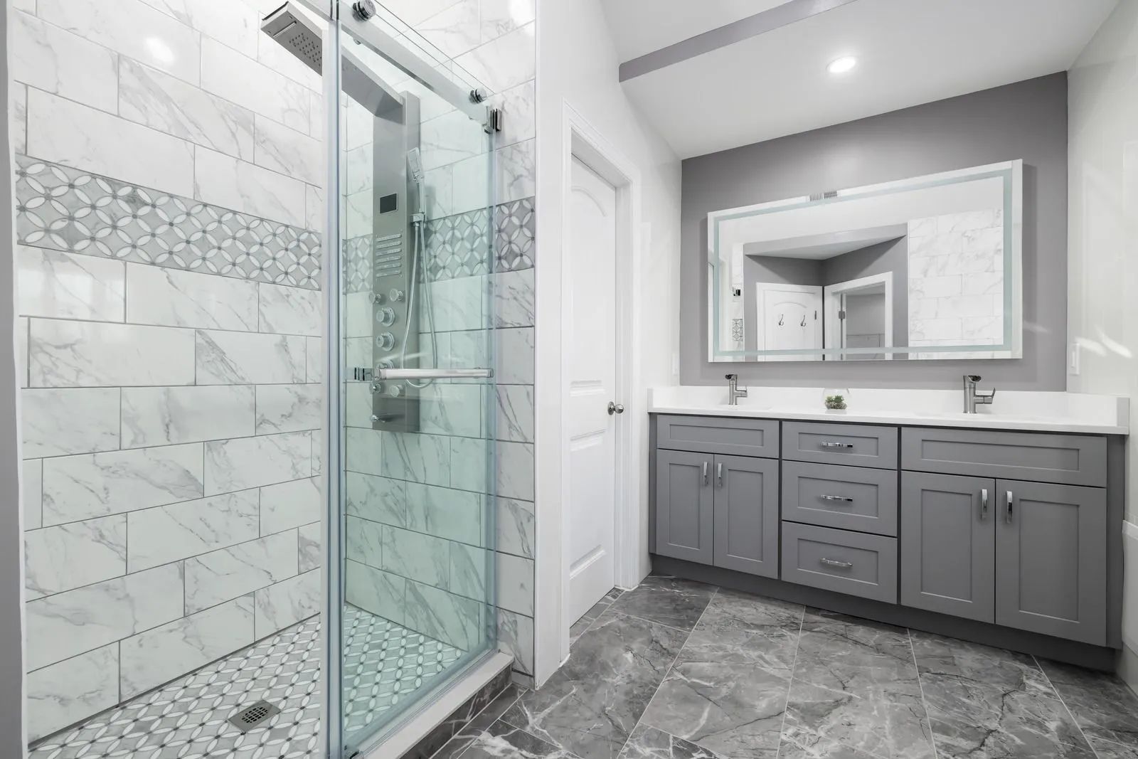 A modern bathroom with a glass-enclosed shower, grey vanity cabinets, a white countertop, and patterned tile flooring.