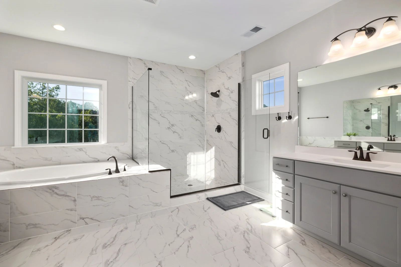 Spacious modern bathroom with marble tiles, glass shower, soaking tub, gray vanity, and large window.