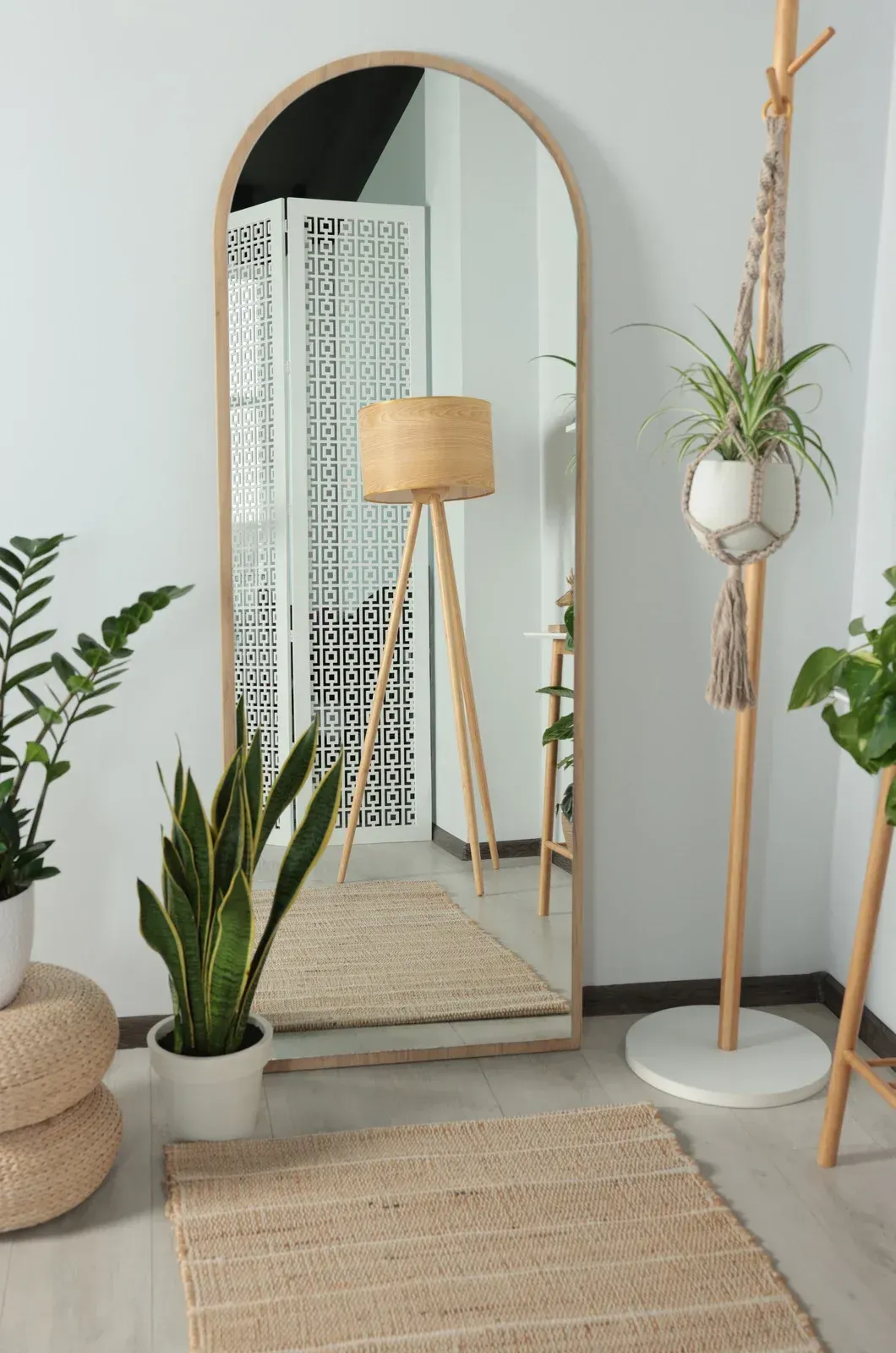 A tall, arched mirror reflects a room with plants, a floor lamp, and a woven rug, creating a bright, natural setting.
