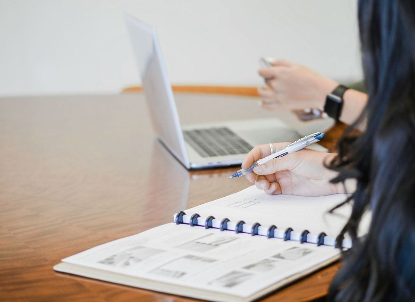 A woman is writing in a notebook while using a laptop computer.