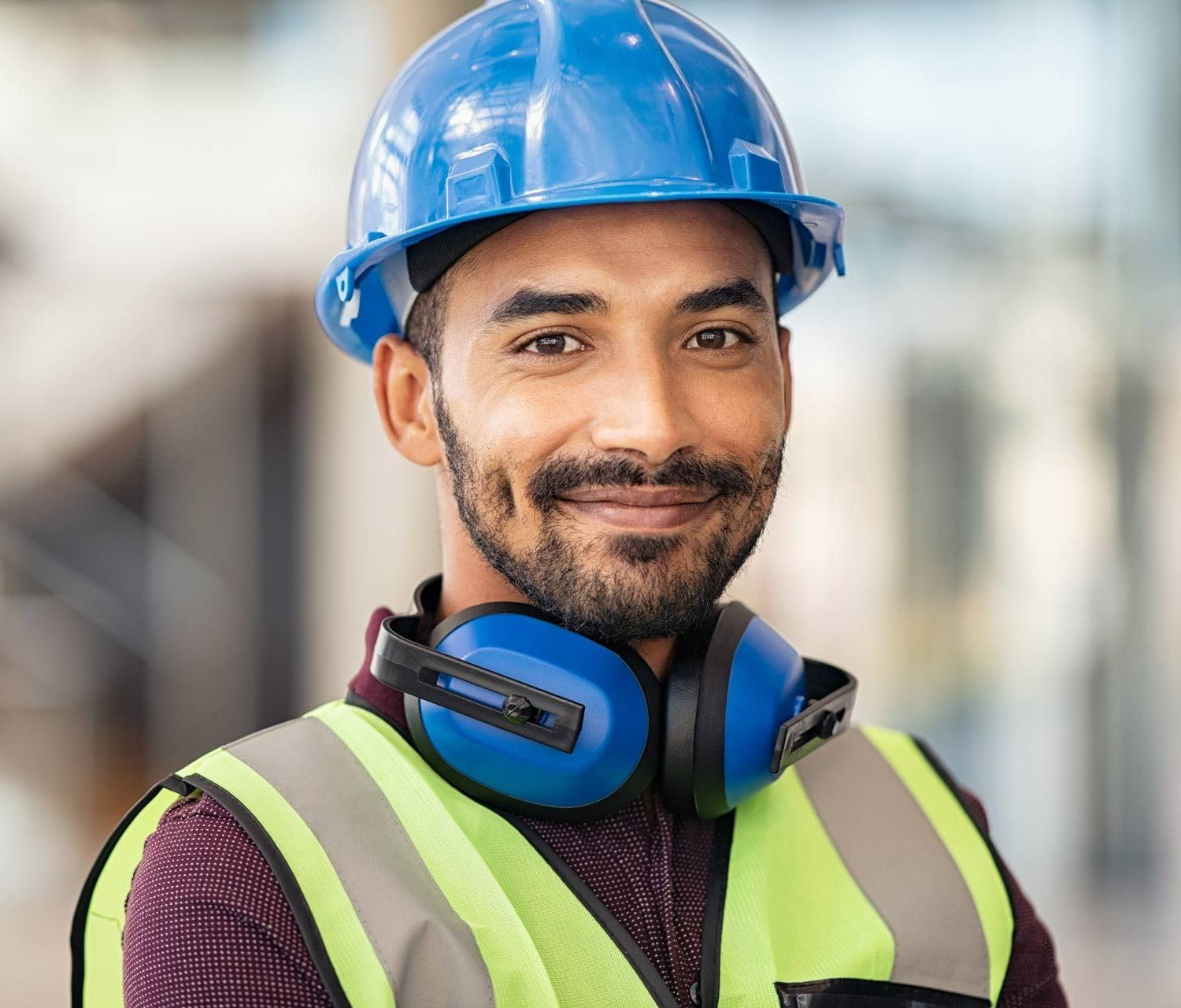Construction worker with a blue hard hat, safety vest, and ear protection, smiling.