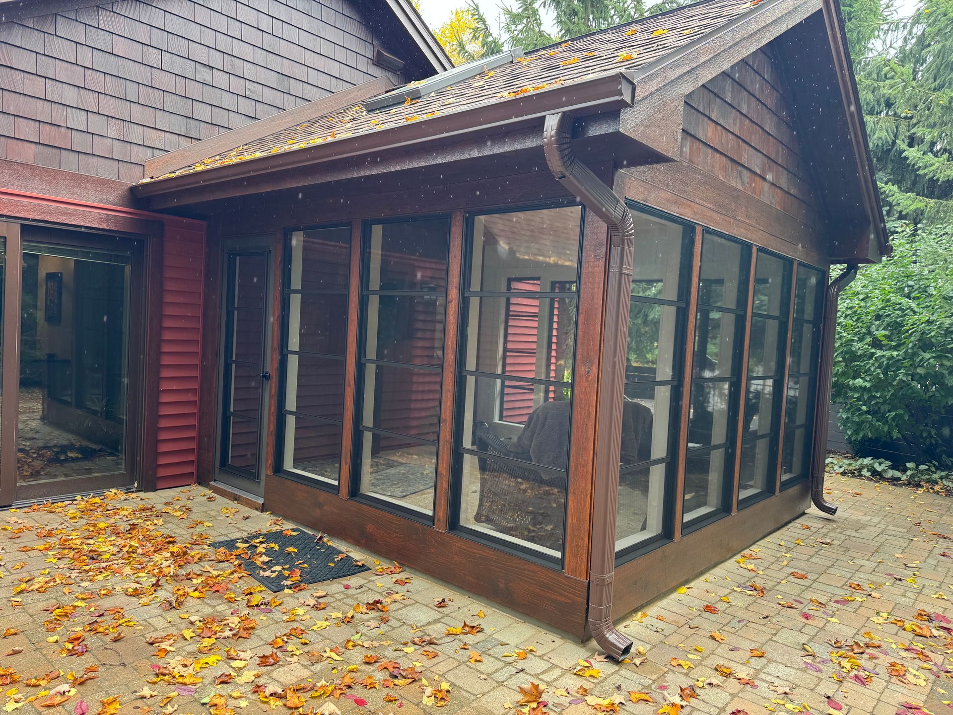 A brown wood-paneled sunroom with large windows, attached to a brick-paved patio with fallen leaves.
