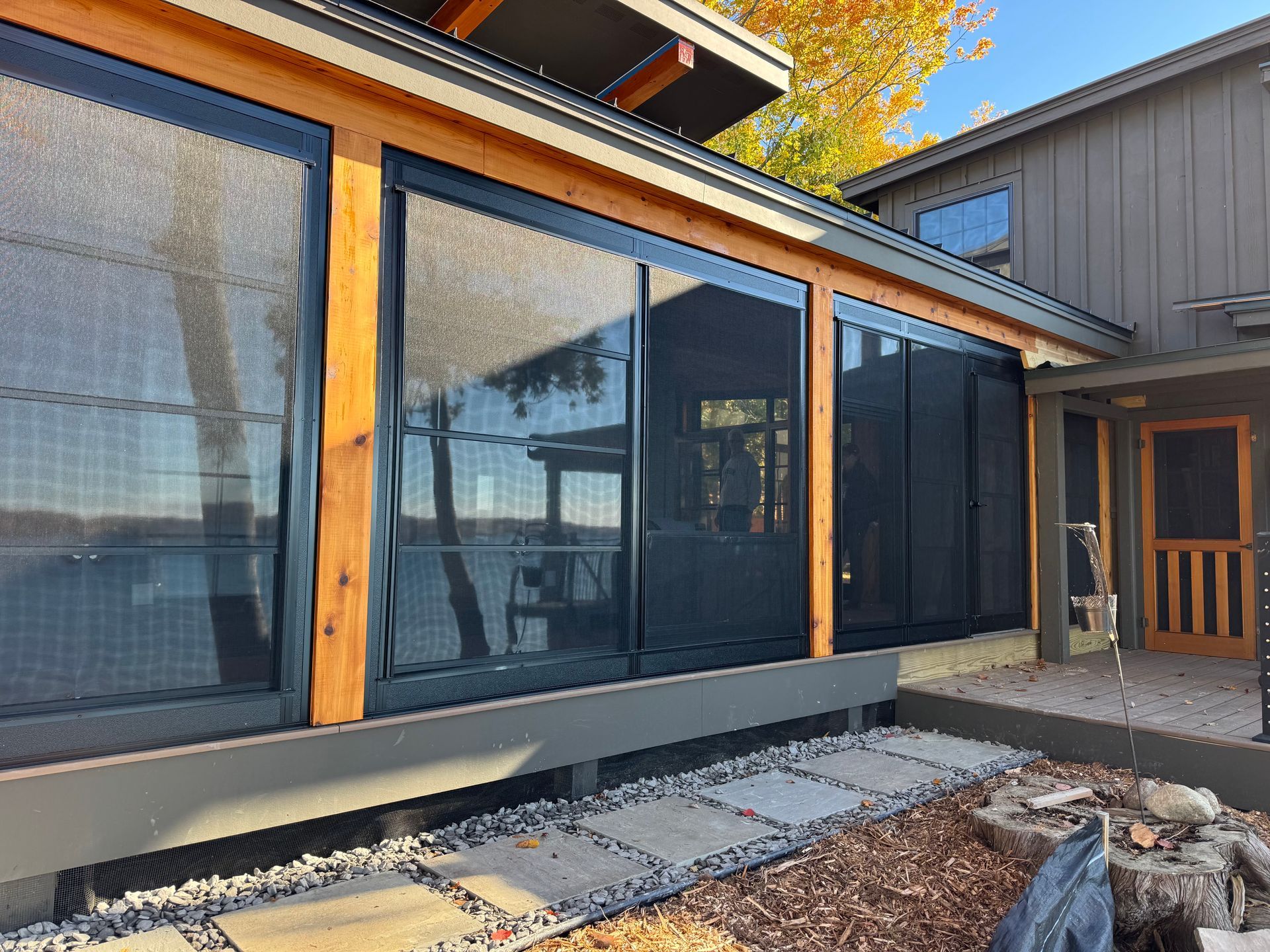 A porch with black screen windows overlooking a lake. The porch has a wooden frame, a stone walkway and a wooden door.