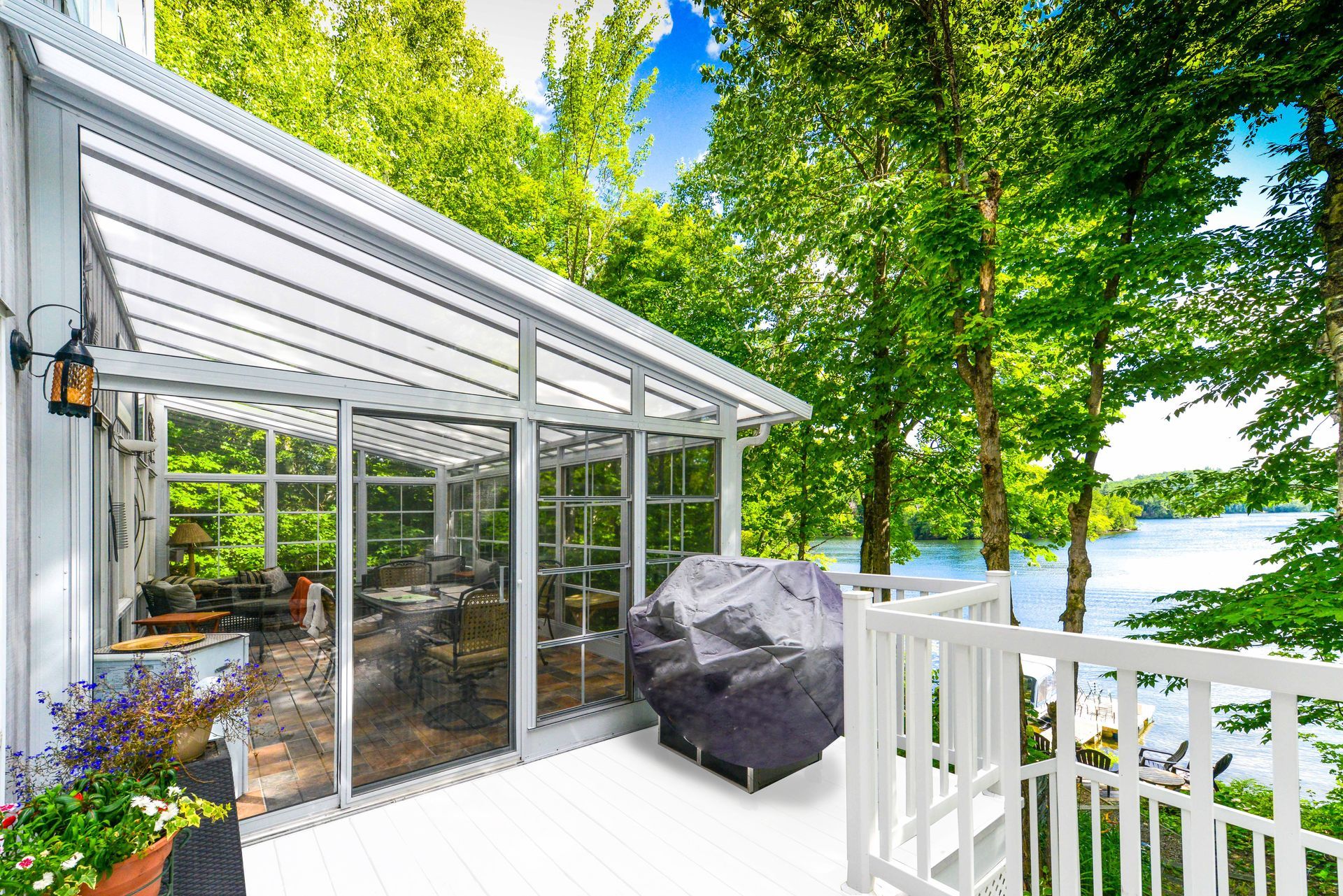 Sunroom attached to a house with a grill and lake view. White deck railing and trees.