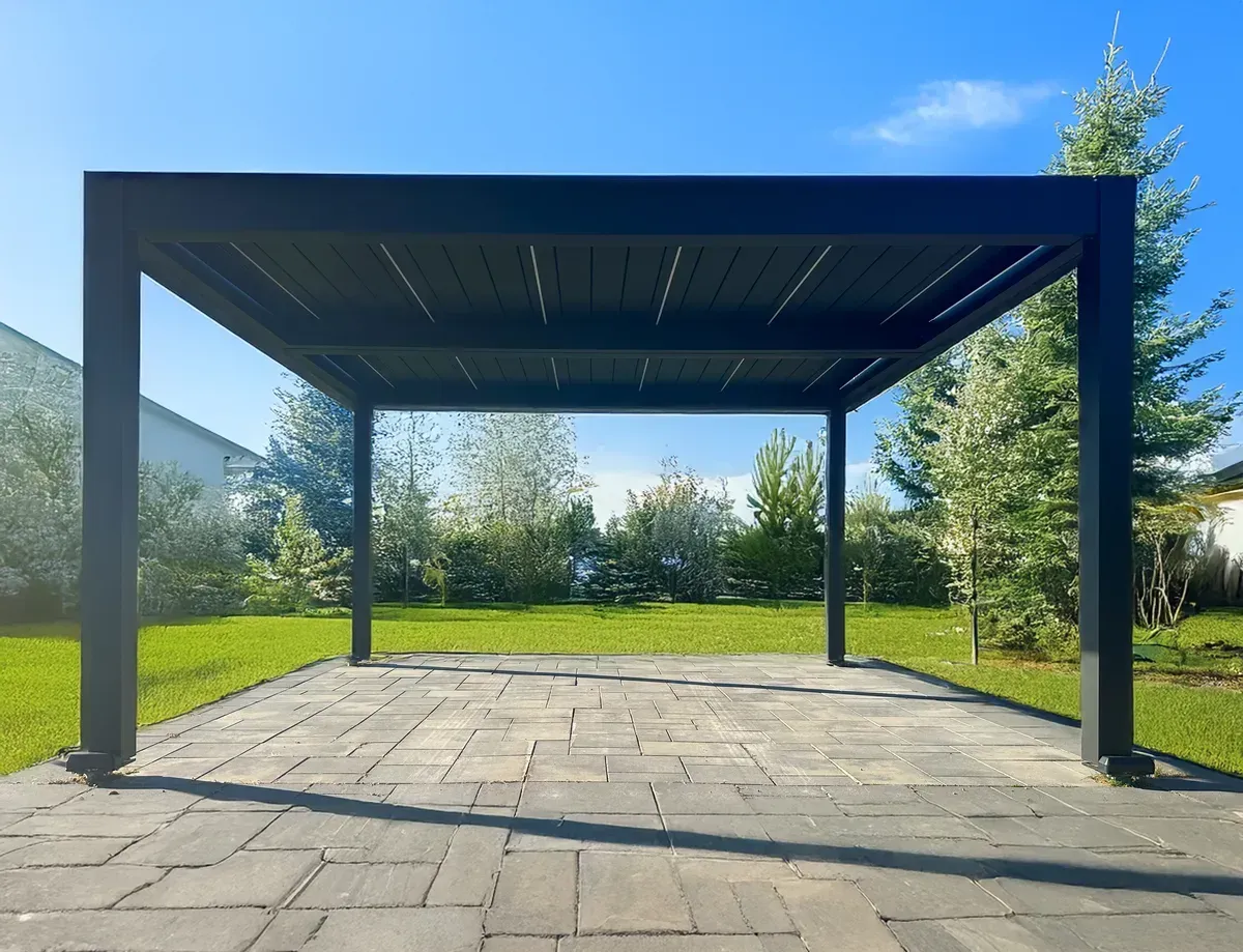 Black pergola on a brick patio with a green lawn and trees in the background under a blue sky.